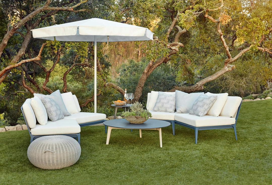 Still modular collection with white fabric, white and grey pattern pillows, and blue finish with round coffee table, side table, and umbrella on a bright green lawn and forest in the background. There's a round Juno ottoman on the side.